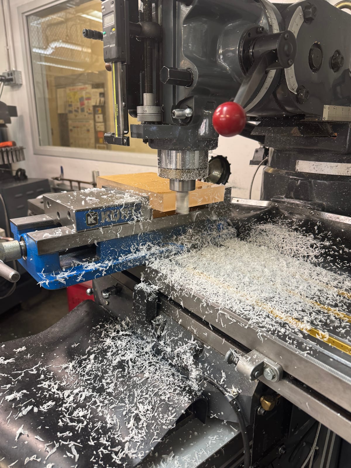 A lathe turning down aluminium bar stock held in a chuck, a stream of curled white aluminium chips coming off the cut, with the machine's digital readout and cross-slide dials visible.