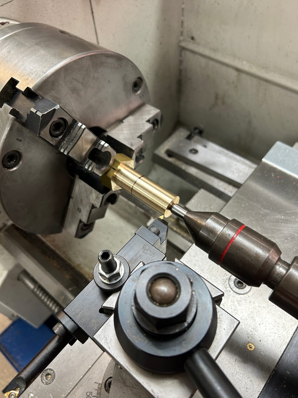 A brass part held in a three-jaw chuck on a mill, an end mill descending toward it, with a hold-down clamp and an edge finder ball visible nearby.