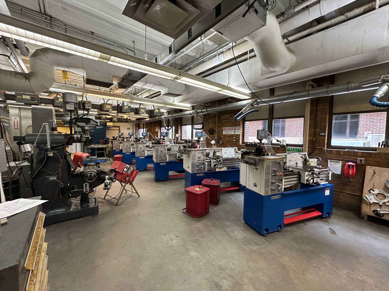A row of engine lathes on blue and red stands running down one side of a university machine shop, with milling machines along the far wall and brick windows behind.