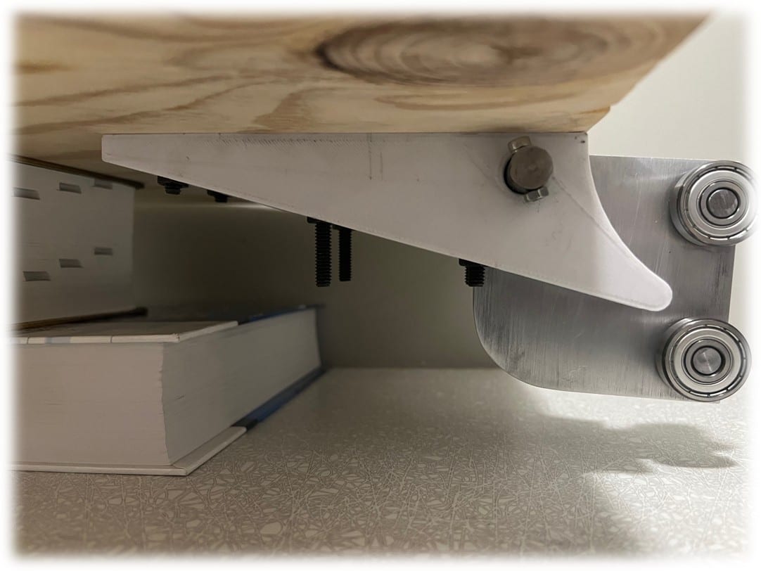 A close-up from underneath the deployed workstation showing a machined aluminium carriage bracket with two ball bearings on the right, bolts hanging through the plywood surface, and a thick book used to prop the assembly for the photo.