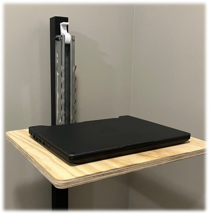 A prototype folding workstation clamped to a vertical black column against a wall, its plywood work surface extended horizontally with a laptop resting on it, a machined steel carriage and hinge visible where the surface meets the column.
