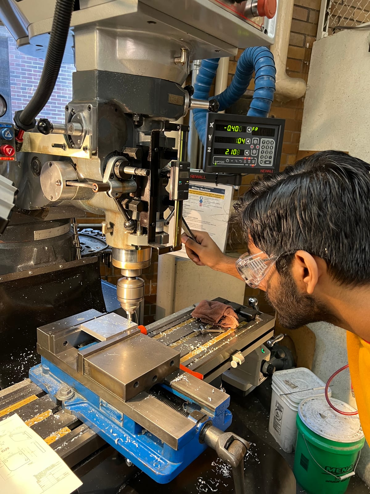 A person in safety glasses working at a manual milling machine with a digital readout, machining a metal part held in a blue vise, aluminium chips on the machine bed.