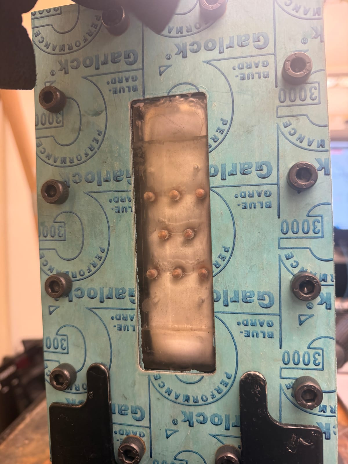 Top-down view of the assembled conductive-gap membrane distillation module during a test: a teal Garlock Blue-Gard gasket bolted around a rectangular window that exposes the membrane, with the pin-fin bumps and beads of permeate condensation visible through it.